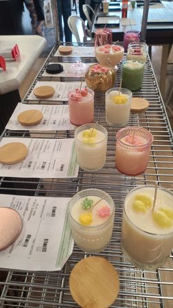 Artisan handmade candles in colorful glass jars with floral wax toppers and bamboo lids arranged on a metal shelving rack with printed labels — craft studio display.