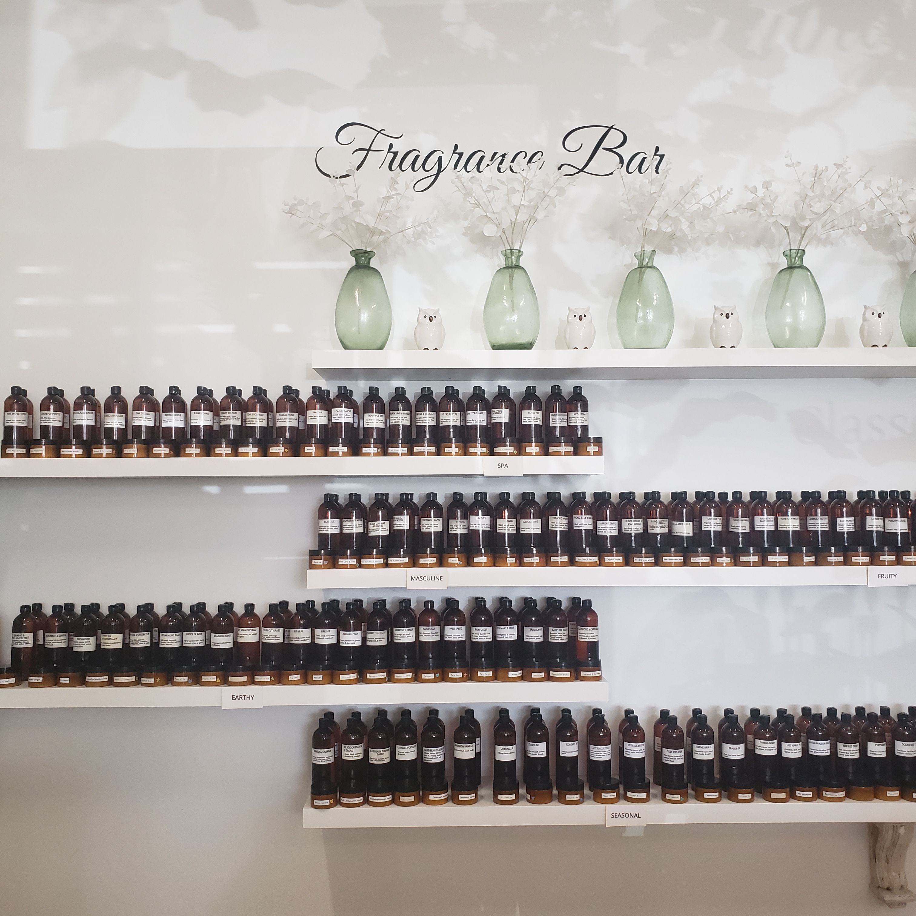 Fragrance bar display with rows of amber perfume sample bottles on white floating shelves labeled by scent categories, accented by green glass vases and white flowers.