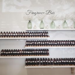 Fragrance bar display with rows of amber perfume sample bottles on white floating shelves labeled by scent categories, accented by green glass vases and white flowers.
