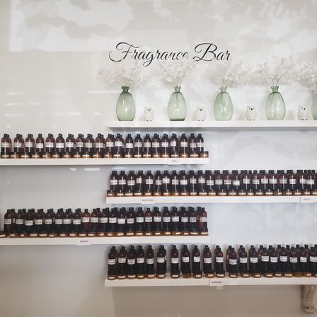Fragrance bar display with rows of amber perfume sample bottles on white floating shelves labeled by scent categories, accented by green glass vases and white flowers.