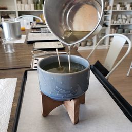 Green wax being poured into a blue ceramic vessel with two wicks set on a small wooden tripod in a candle-making studio workshop, worktable and shelves visible in the background
