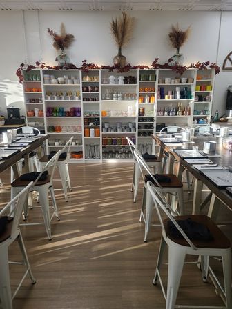 Bright, cozy craft workshop interior with long wooden tables and white metal bar stools set for a class, wall of shelves filled with colorful glass jars, candles and craft supplies, autumn garland and dried floral accents on top, sunlight streaming across the wood floor.