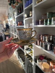Hand holding an ornate gold-trimmed porcelain teacup and saucer in a bright home decor boutique aisle lined with candles, jars and vases.