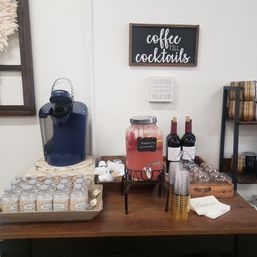 Self-serve beverage station on wooden table: blue Keurig coffee maker, glass dispenser labeled Raspberry Lemonade, bottled water, two wine bottles, stacked plastic and gold cups and napkins beneath a 'coffee till cocktails' wall sign.