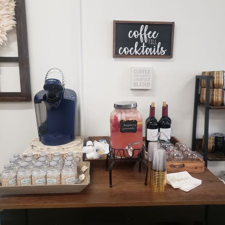 Self-serve beverage station on wooden table: blue Keurig coffee maker, glass dispenser labeled Raspberry Lemonade, bottled water, two wine bottles, stacked plastic and gold cups and napkins beneath a 'coffee till cocktails' wall sign.