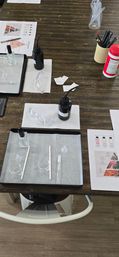 Overhead view of a wooden workshop table set for perfume-making: trays with glass vials, small beakers, droppers and funnels, scent strips and pencils, safety goggles, black squeeze bottles and printed scent charts, ready to mix.