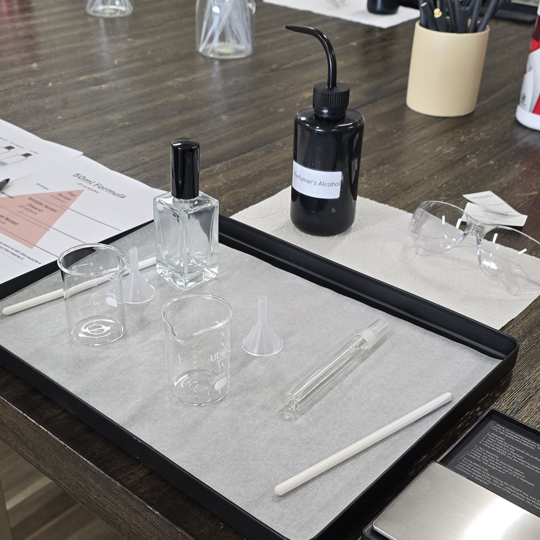 Fragrance workshop setup on a dark wood table: clear glass perfume bottle, small glass beakers and funnels, plastic spray vial, stirring rod, a labeled bottle of perfumer's alcohol and safety glasses for DIY perfume blending.