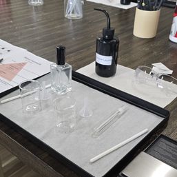 Fragrance workshop setup on a dark wood table: clear glass perfume bottle, small glass beakers and funnels, plastic spray vial, stirring rod, a labeled bottle of perfumer's alcohol and safety glasses for DIY perfume blending.