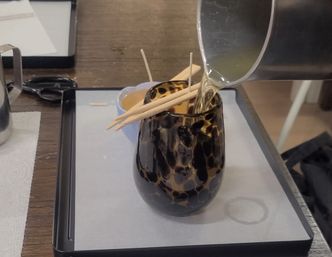 Pouring hot wax into a tortoiseshell-patterned glass votive as wooden skewers hold wicks in place during candle-making on a prep tray.