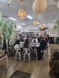 Group fragrance workshop in a cozy boutique studio — attendees seated at a communal table sampling scents, rows of bottles on shelves, rattan pendant lights and hanging greenery.