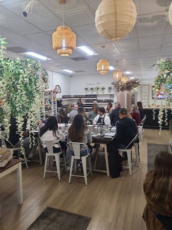 Group fragrance workshop in a cozy boutique studio — attendees seated at a communal table sampling scents, rows of bottles on shelves, rattan pendant lights and hanging greenery.