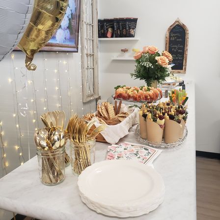 Indoor party buffet on a marble table with gold utensils in jars, stacked white plates, breadsticks and crackers, shrimp cocktail platter, paper cones of cheese-and-fruit skewers, peach rose bouquet and twinkling fairy lights.