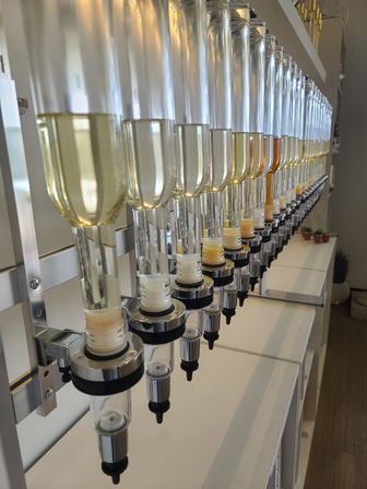 Row of wall-mounted clear dispensing bottles with measured pour spouts holding pale yellow to amber liquids above a white countertop — modern tasting station interior.