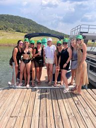 Ten friends posing on a pontoon boat at a lakeside dock, wearing swimsuits and matching green caps with one in a white bride hat, holding drinks with wooded hills and a partly cloudy summer sky in the background.