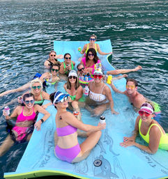 Group of friends in colorful swimwear lounging on a large blue floating mat in a lake, holding canned drinks and smiling at a fun summer water party.
