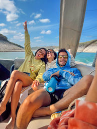 Three friends laughing on a sunny lake boat ride, wearing hoodies and sunglasses with blue sky and rocky shoreline