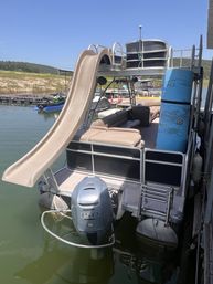 Pontoon boat docked on a sunny lake featuring a tan water slide, cushioned seating, blue foam tube and 150 HP outboard motor, with jet skis and shoreline in the background.