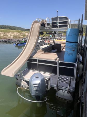 Pontoon boat docked on a sunny lake featuring a tan water slide, cushioned seating, blue foam tube and 150 HP outboard motor, with jet skis and shoreline in the background.