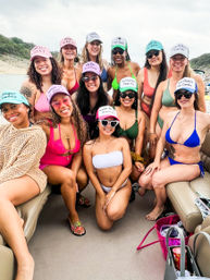 Group of smiling women on a lake boat in colorful bikinis and novelty trucker hats, wearing sunglasses and posing together near a rocky shoreline on an overcast day — fun girls' boat party