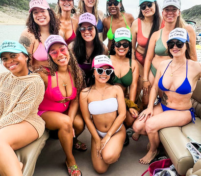 Group of smiling women on a lake boat in colorful bikinis and novelty trucker hats, wearing sunglasses and posing together near a rocky shoreline on an overcast day — fun girls' boat party