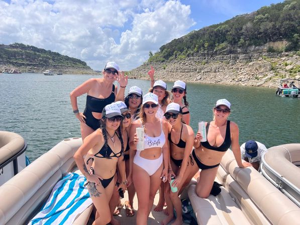 Happy group of women in swimsuits and matching caps celebrating a bachelorette on a pontoon boat on a sunny lake with rocky cliffs