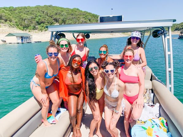 Ten friends in colorful swimsuits and sunglasses smiling and posing on a pontoon boat at a sunny lake with a green wooded shoreline