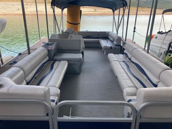 Spacious pontoon boat interior with gray-and-blue cushioned bench seating, covered canopy, and open central deck docked at a calm lake shore.