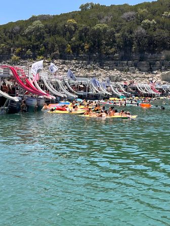 Crowded summer lake scene with party boats and pontoons lined at a dock, inflatable water slides and flags, dozens of people on colorful floaties in clear turquoise water by a rocky, tree-lined shoreline.