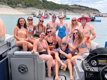 Group of friends enjoying a summer lake party on a pontoon boat, wearing swimsuits and sunglasses, holding drinks and smiling with anchored boats and a rocky shoreline in the background.