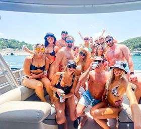 Smiling group of young adults in swimsuits enjoying a sunny summer boat party on a lake, holding drinks with wooded hills and a dock in the background.