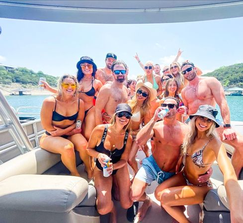 Smiling group of young adults in swimsuits enjoying a sunny summer boat party on a lake, holding drinks with wooded hills and a dock in the background.