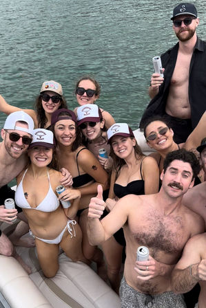 Smiling group of friends at a summer boat party on a lake, wearing swimsuits and trucker hats, holding canned drinks aboard a crowded pontoon — upbeat, sun-soaked vibe.