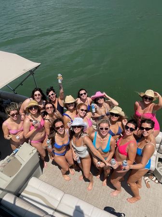 Pontoon boat party on a lake with a group of women in colorful bikinis and sun hats, holding canned drinks and smiling toward the camera on a sunny summer day.