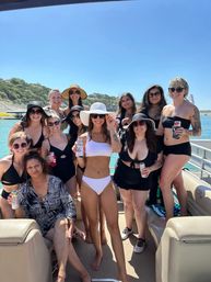 Group of women in swimsuits and sun hats on a pontoon boat enjoying a sunny lake day, holding canned drinks with wooded shoreline and clear blue sky in the background.