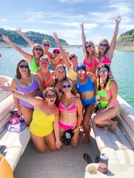 Sun-soaked lake day: a cheerful group of women in bright, colorful swimsuits and sunglasses posing and cheering on a pontoon boat with rocky, tree-lined shores in the background.
