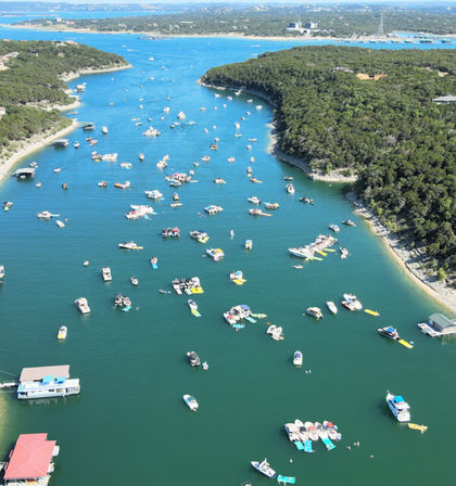 Aerial view of a busy summer lake cove with dozens of boats, pontoons, and colorful floating platforms clustered in blue-green water along a tree-lined shoreline.