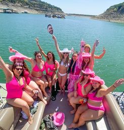 Group of women in bright pink swimsuits and fuzzy cowboy hats partying on a pontoon boat at a sunny lake, raising drinks and a face-cutout paddle with rocky shoreline and other boats in the background.