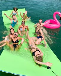 Group of friends laughing and splashing on a bright green floating mat in a sunny lake, wearing swimsuits and sunglasses beside a pink inflatable flamingo.