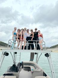 Eight friends in swimsuits smiling on the upper deck of a pontoon boat over a lake with cloudy sky and tree-lined shoreline, summer boat outing