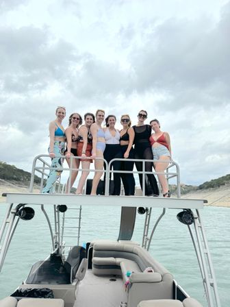 Eight friends in swimsuits smiling on the upper deck of a pontoon boat over a lake with cloudy sky and tree-lined shoreline, summer boat outing