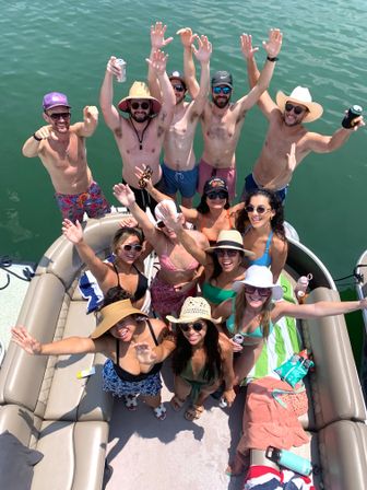 Aerial view of a group of friends waving on a pontoon boat on a sunny lake day, wearing swimsuits, hats and sunglasses and holding drinks.