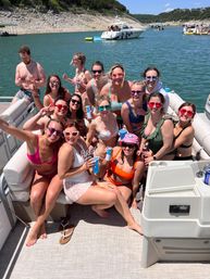 Smiling group of friends on a pontoon boat lake party—women in colorful swimsuits and sunglasses holding drinks on a sunny summer day with other boats and a rocky shoreline in the background.