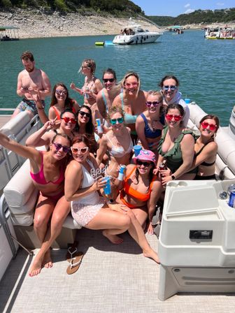 Smiling group of friends on a pontoon boat lake party—women in colorful swimsuits and sunglasses holding drinks on a sunny summer day with other boats and a rocky shoreline in the background.