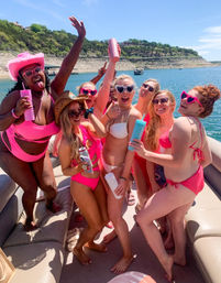 Group of women on a sunny pontoon boat for a lake day, wearing pink swimsuits and heart-shaped sunglasses, laughing and holding colorful drink tumblers with blue water and rocky shoreline behind them.