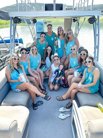 Group of women in matching teal tank tops and heart-shaped sunglasses posing with drinks and a novelty groom doll on a pontoon boat at a sunny lake