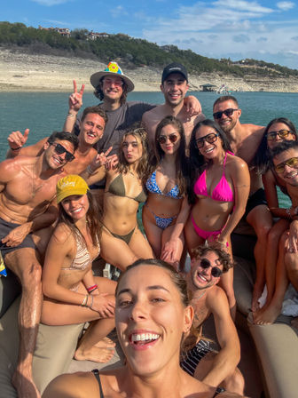 Sun-soaked group of friends in swimsuits taking a selfie on a boat at a scenic lake with a rocky shoreline and hillside homes in the background