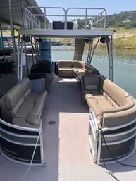 Spacious pontoon boat interior with tan cushioned wraparound seating, upper deck with speakers, docked at a sunny lake marina