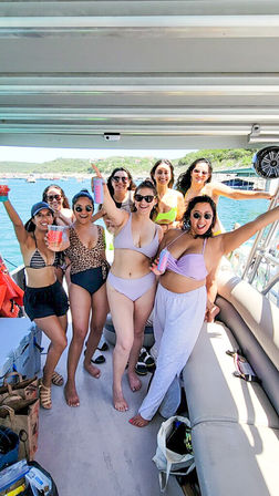 Smiling group of women in swimsuits cheering with drinks on a pontoon boat during a sunny lake day with green hills and other boats in the background.