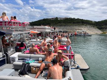 Crowded lake party with linked pontoon boats and groups of friends in swimsuits sunbathing and drinking, pink boat seating, rocky shoreline and hilltop homes under a partly cloudy sky