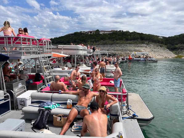 Crowded lake party with linked pontoon boats and groups of friends in swimsuits sunbathing and drinking, pink boat seating, rocky shoreline and hilltop homes under a partly cloudy sky